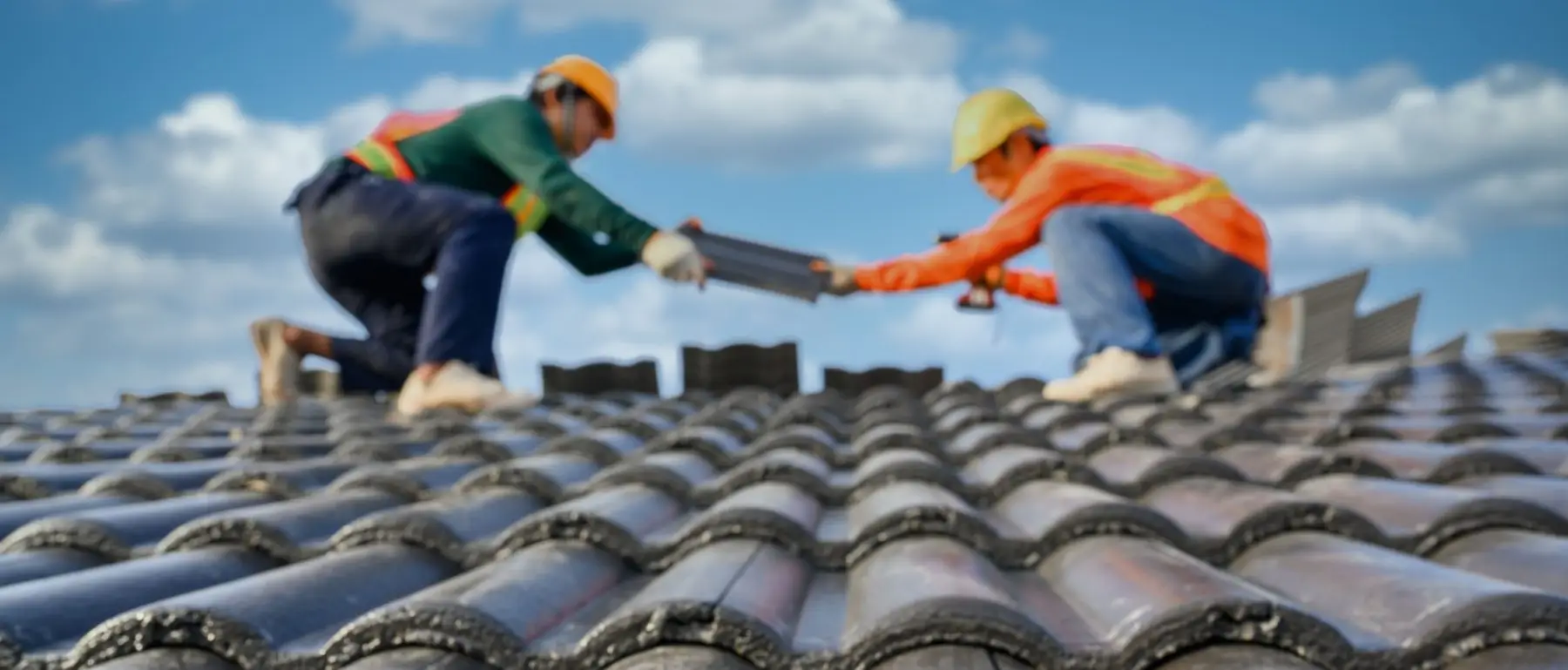 Two workers in safety gear passing roofing tiles on a sunny day.